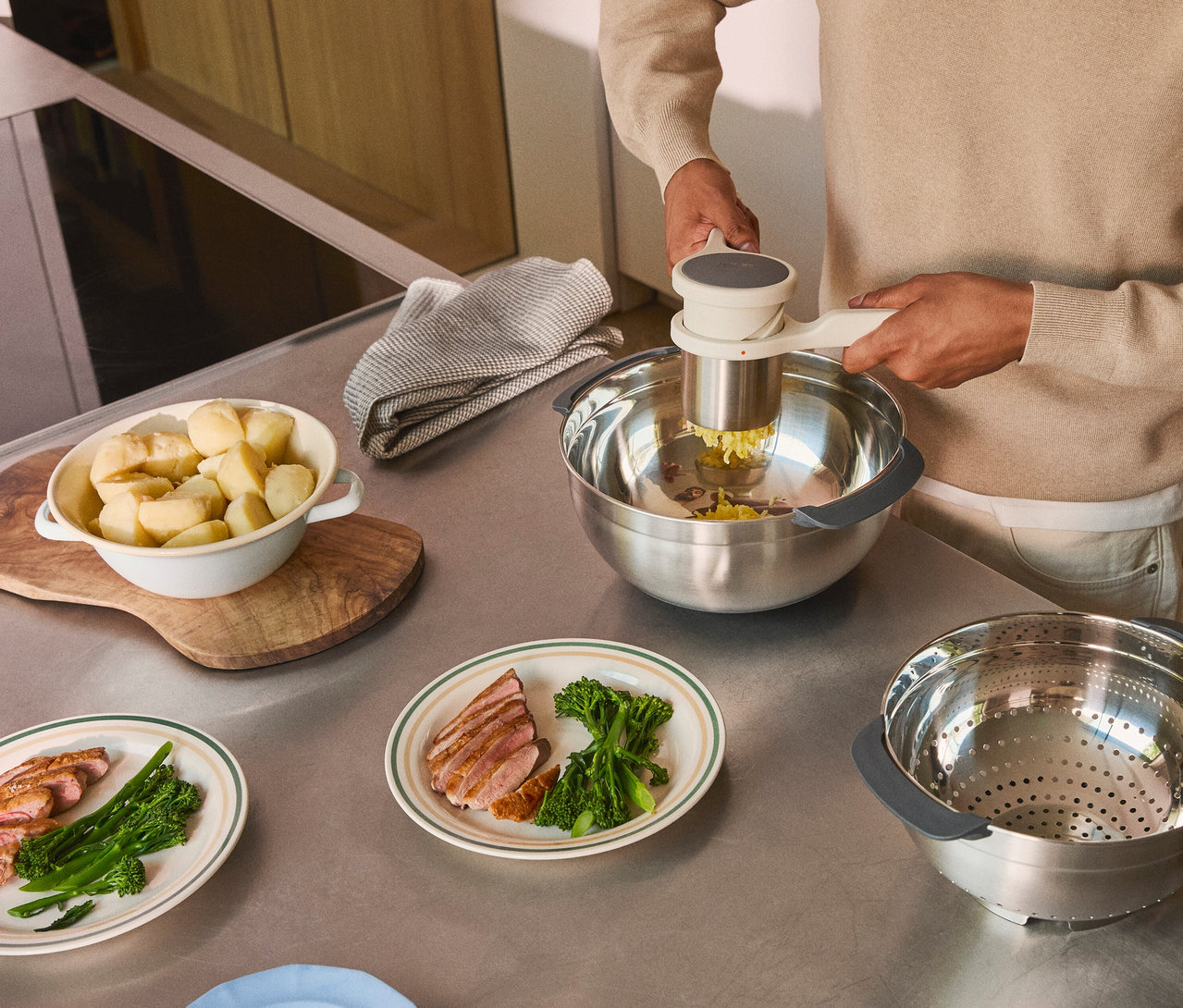 Mann zerdrückt Kartoffeln in einer Edelstahlschüssel mit einem Kartoffelstampfer. Auf dem Tisch liegen Teller mit Fleisch und Brokkoli.