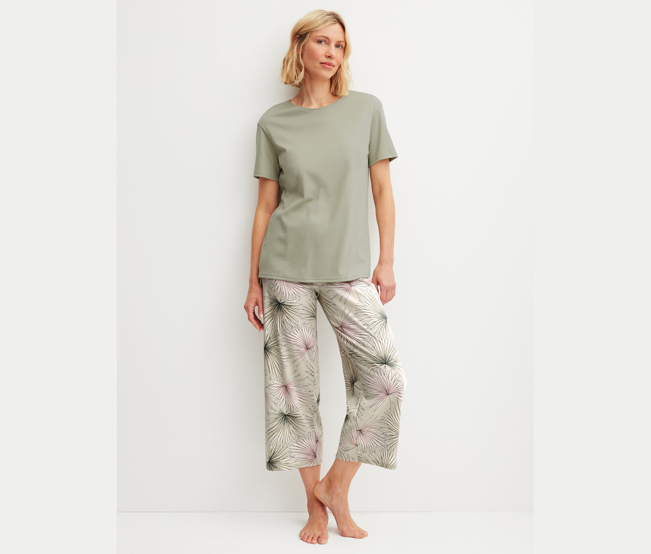 Eine Frau mit blonden Haaren trägt ein olivfarbenes T-Shirt und gemusterte Hosen.