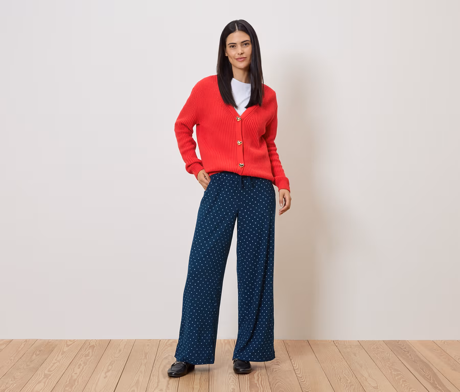 Frau mit roter Strickjacke und blau-weiß gepunkteter Hose steht auf Holzboden vor einer Wand.