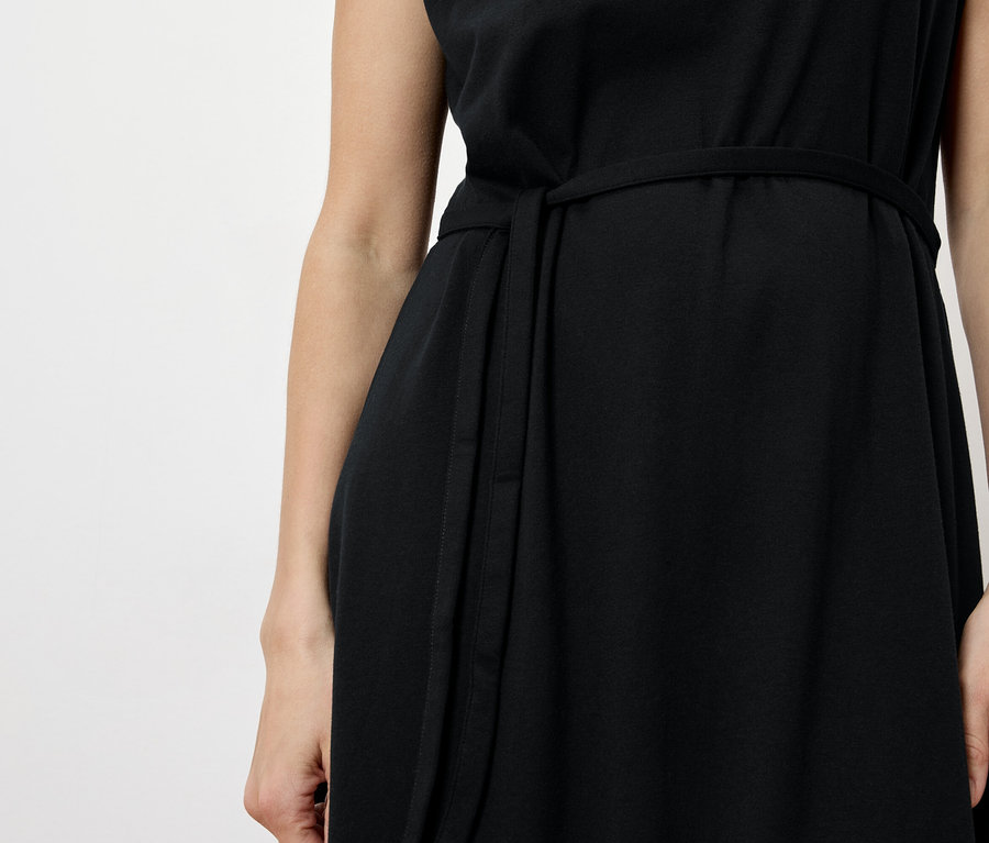 Nahaufnahme einer Frau in einem schwarzen Kleid mit Gürtel vor einem weißen Hintergrund.