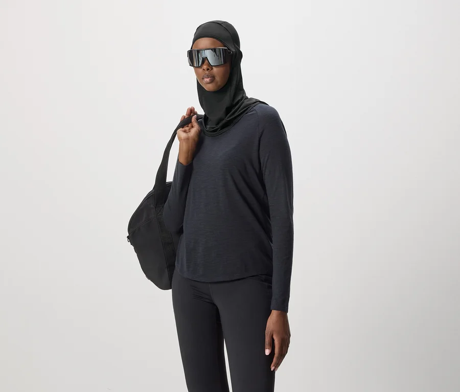 Frau mit Hijab, Sonnenbrille, schwarzem Oberteil und Leggings, mit schwarzer Tasche über der Schulter vor neutralem Hintergrund.