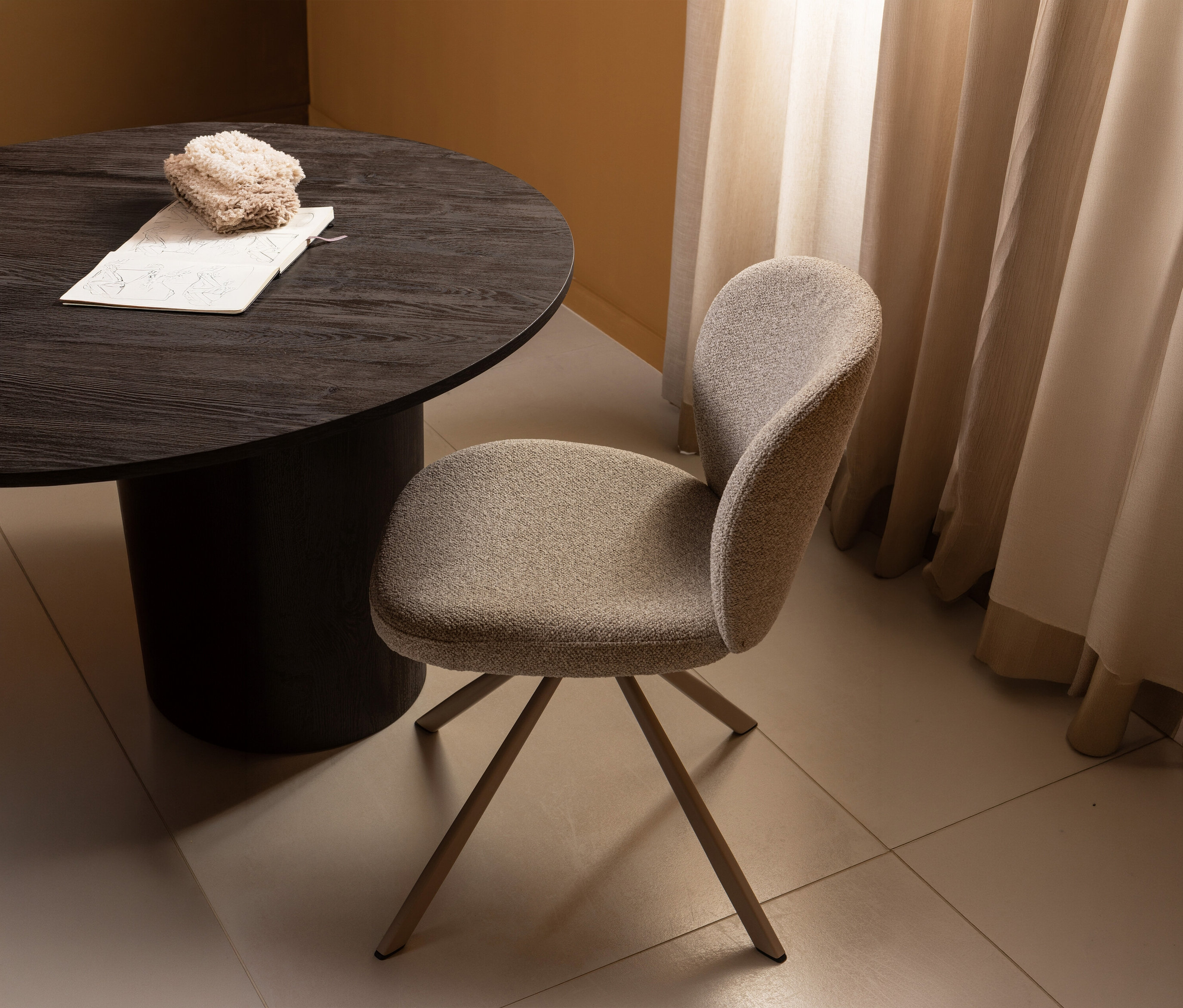 Ein WOOOD »Femke« Stuhl aus einem 2er-Set steht an einem runden, dunklen Tisch mit Notizbuch und beiger Decke vor einem beigen Vorhang.
