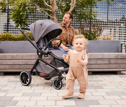 Kleinkind in beigem Overall steht vor grauem Kinderwagen, Mann sitzt auf Bank im Hintergrund.