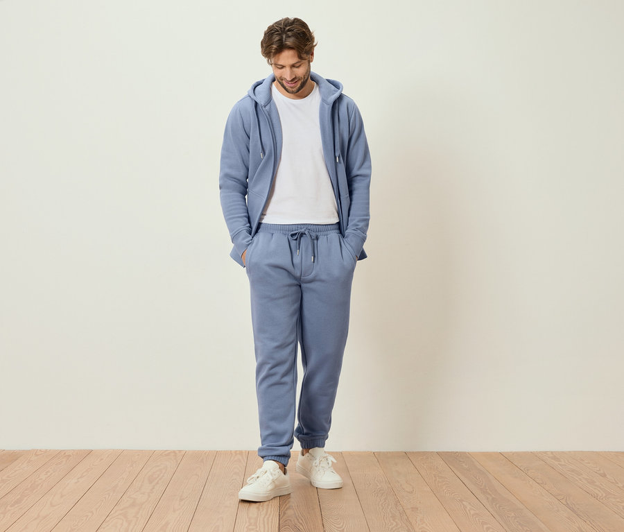 Mann steht mit Händen in den Taschen in hellblauem Trainingsanzug und weißen Turnschuhen.