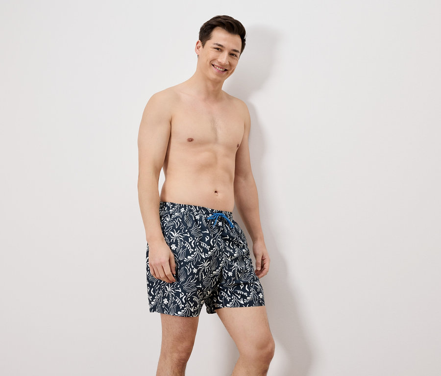 Ein Mann mit Badehose steht vor einer weißen Wand.