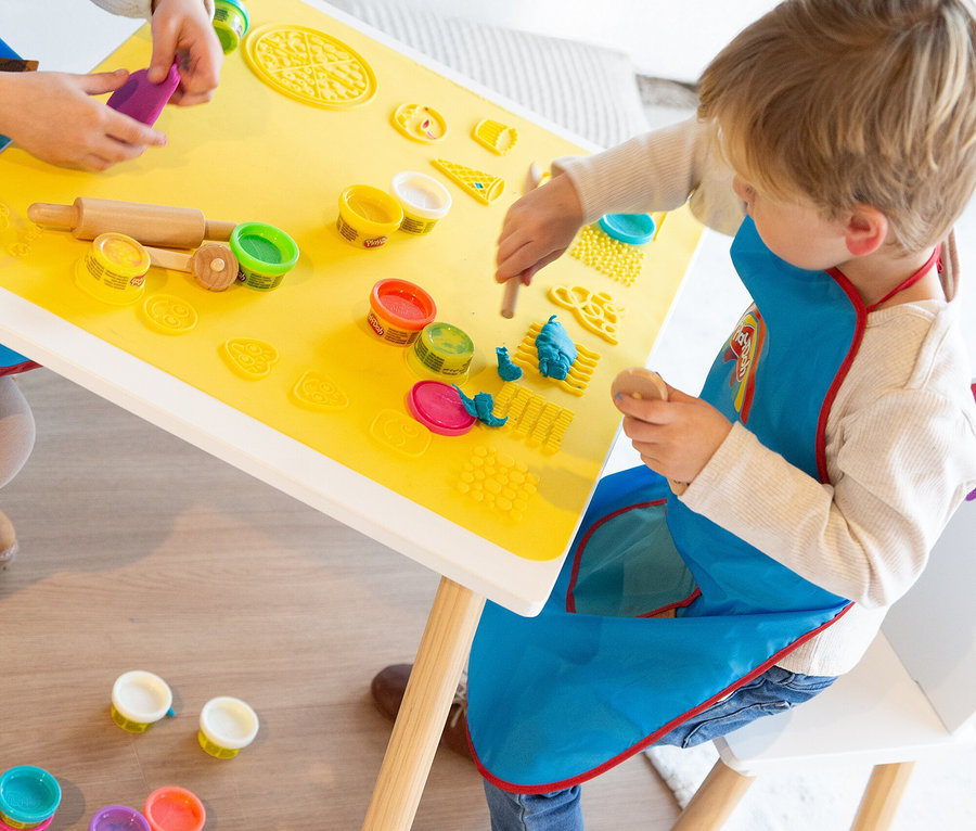 Zwei Kinder sitzen an einem gelben Tisch und spielen mit Knete. Auf dem Tisch liegen Knetdosen, eine Teigrolle und Förmchen.