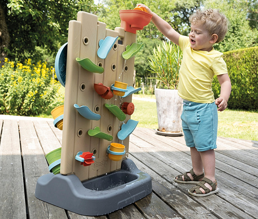 Junge gießt Wasser in die Smoby Life Activity Wall. Das Wasser läuft an bunten Elementen herunter in ein Becken.