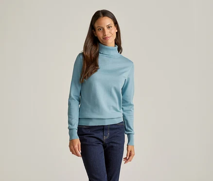 Frau mit langen braunen Haaren, die einen hellblauen Rollkragenpullover und eine dunkle Jeans vor einem hellen Hintergrund trägt.