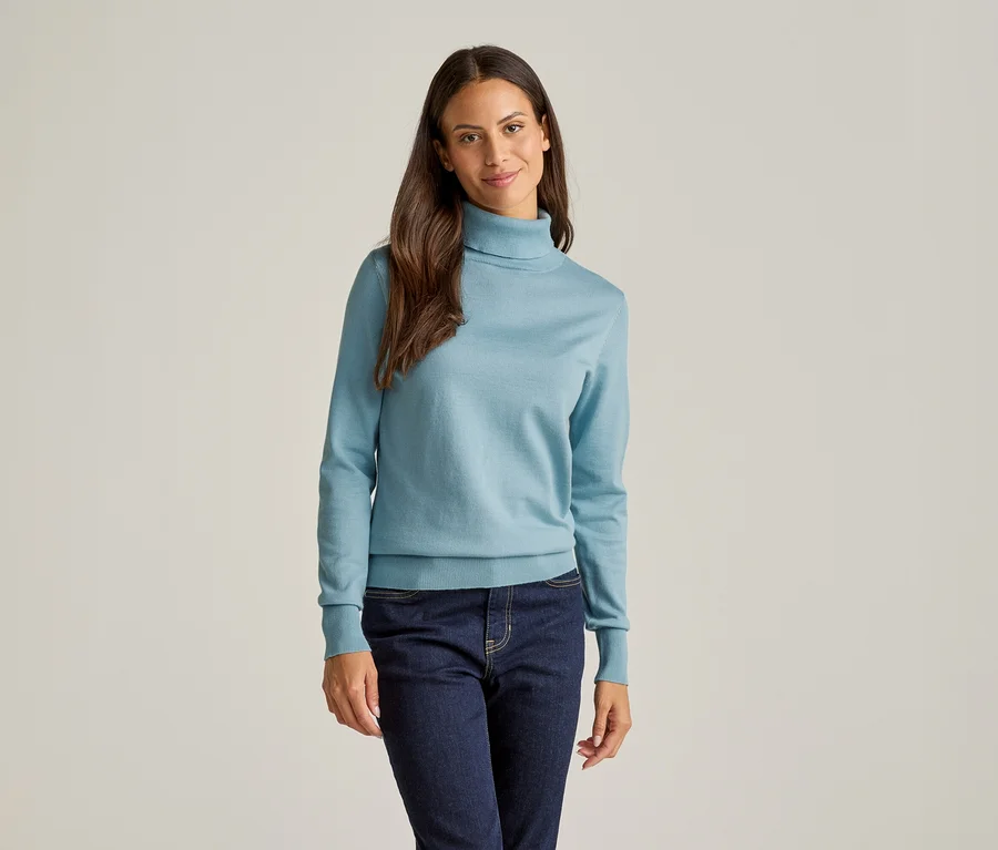Frau mit langen braunen Haaren, die einen hellblauen Rollkragenpullover und eine dunkle Jeans vor einem hellen Hintergrund trägt.