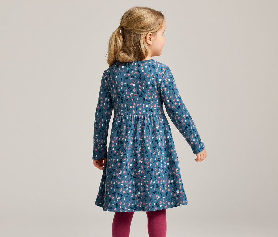 Von hinten ist ein Mädchen mit blonden Haaren in einem blauem, langärmligen Kleid mit Blumenmuster und Leggings zu sehen.