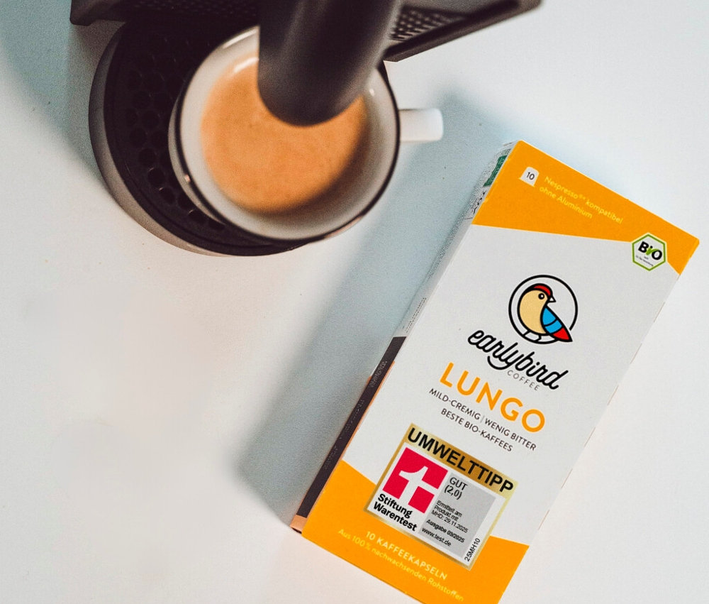 Eine Tasse Kaffee unter einer Kaffeemaschine und eine Packung Earlybird Lungo Kaffee sind auf einem Tisch zu sehen.
