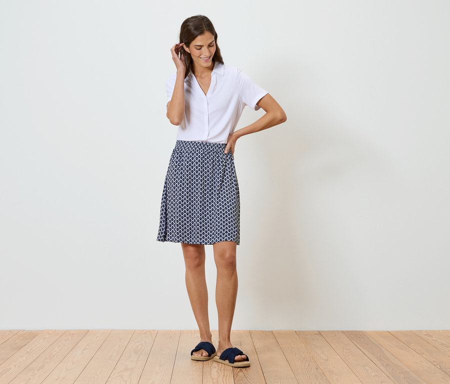 Frau steht vor einer weißen Wand und trägt ein weißes Hemd und einen blau-weiß gemusterten Rock.