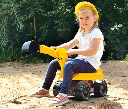 Ein lächelndes Mädchen mit gelbem Schutzhelm sitzt auf einem gelben GIGA TRUCKS Aufsitz-Bagger Pro X im Sand.
