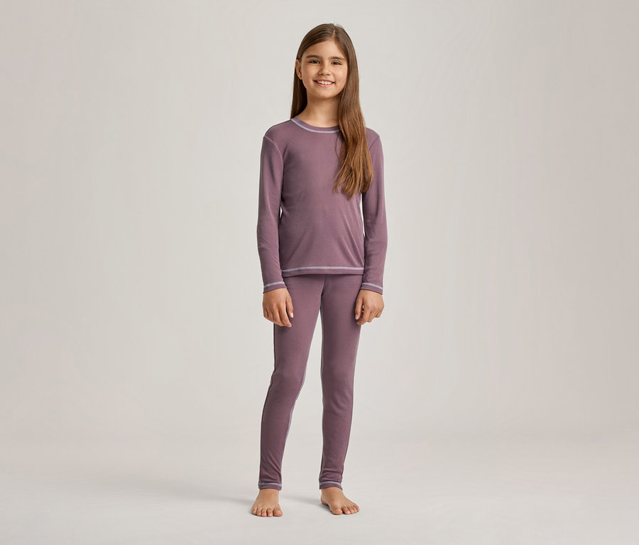 Ein lächelndes Mädchen mit langen braunen Haaren steht barfuß und trägt einen mauvefarbenen Pyjama mit langen Ärmeln und passenden Leggings, die hellgraue Ziernähte haben.