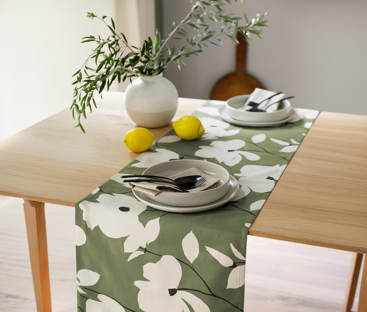 Ein heller Holztisch mit einem SCHÖNER WOHNEN-Kollektion Tischläufer in Khaki mit weißen Blumenmustern. Darauf stehen zwei Gedecke mit grauen Tellern, Schalen und schwarzem Besteck auf Servietten, flankiert von zwei Zitronen. Links oben steht eine Vase mit grünen Zweigen.