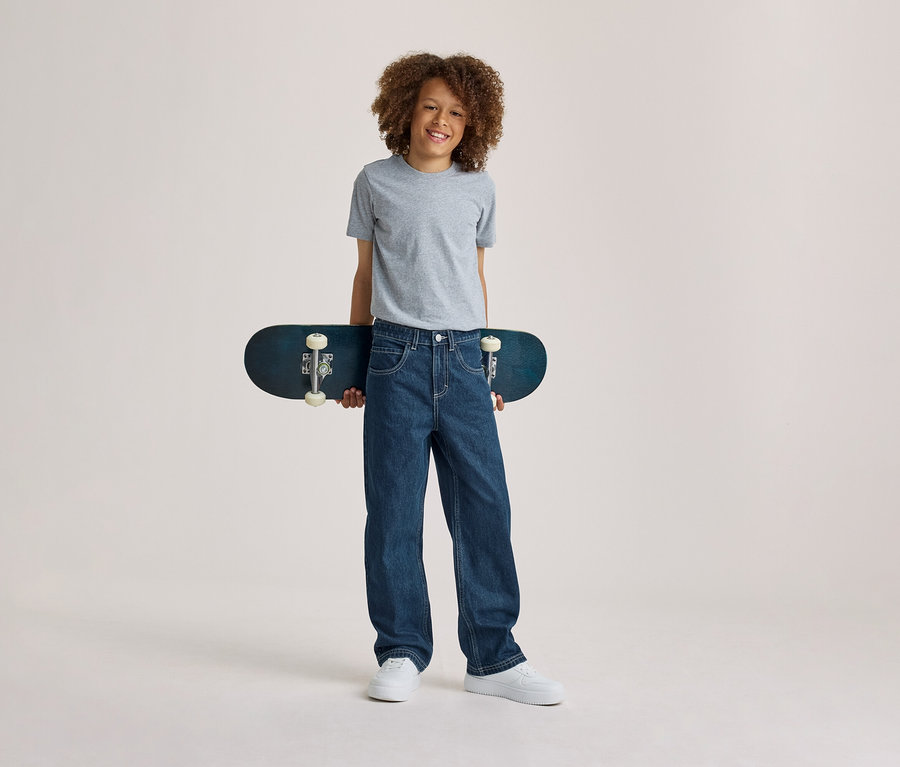 Lächelnder Junge mit lockigem Haar, der ein graues T-Shirt, blaue Jeans und weiße Sneaker trägt und ein dunkelblaues Skateboard hinter seinem Rücken hält.