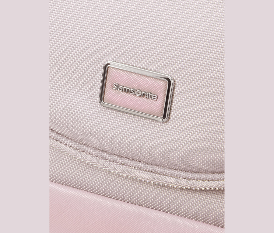 Nahaufnahme einer hellrosa Samsonite Tasche, die ein silbernes rechteckiges Logo-Schild mit „Samsonite“ auf rosa Hintergrund zeigt. Eine geschwungene Naht ist unter dem Logo sichtbar.