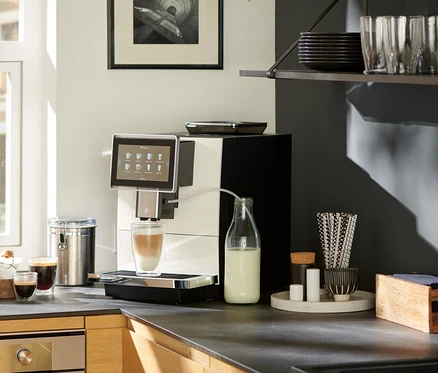 Auf einer Küchentheke steht ein weißer »Tchibo Office« Kaffeevollautomat, der Kaffee in ein Glas zubereitet. Daneben stehen eine Milchflasche, Croissants, Kaffee und ein Notizbuch.