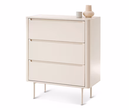 Eine hellbeige Kommode mit drei Schubladen, jeweils mit Griffmulde. Sie hat abgerundete Ecken und steht auf vier dünnen Beinen. Oben stehen ein graues Zylinderobjekt und eine beige bauchige Vase.