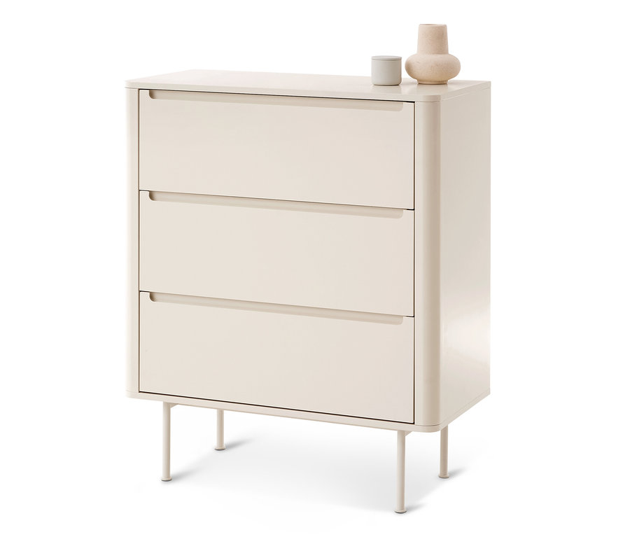 Eine hellbeige Kommode mit drei Schubladen, jeweils mit Griffmulde. Sie hat abgerundete Ecken und steht auf vier dünnen Beinen. Oben stehen ein graues Zylinderobjekt und eine beige bauchige Vase.