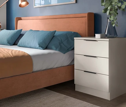 Ein Schlafzimmer mit einem hellbraunen Polsterbett, hellblauen Kissen und einem hellbeigen Nachttisch mit drei Schubladen. Eine kupferfarbene Wandlampe hängt an der dunkelblauen Wand, eine Vase mit Grünpflanzen steht auf dem Nachttisch.