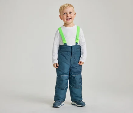 Ein kleiner Junge mit blonden Haaren lächelt in die Kamera. Er trägt ein weißes Langarmshirt, blaue Schneehosen mit grünen Hosenträgern und blau-weiße Turnschuhe. Die Schneehosen haben reflektierende Streifen an Knien und Knöcheln.