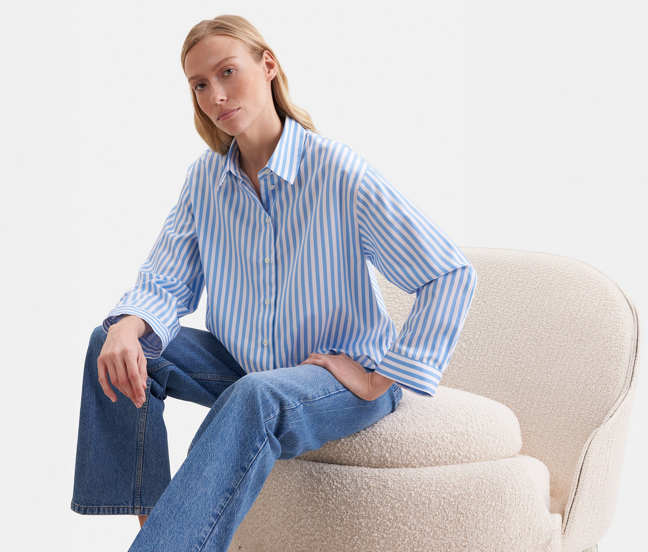Blonde Frau sitzt auf beigem Sessel und trägt eine blau-weiß gestreifte Seidensticker Oversize Hemdbluse und blaue Jeans.