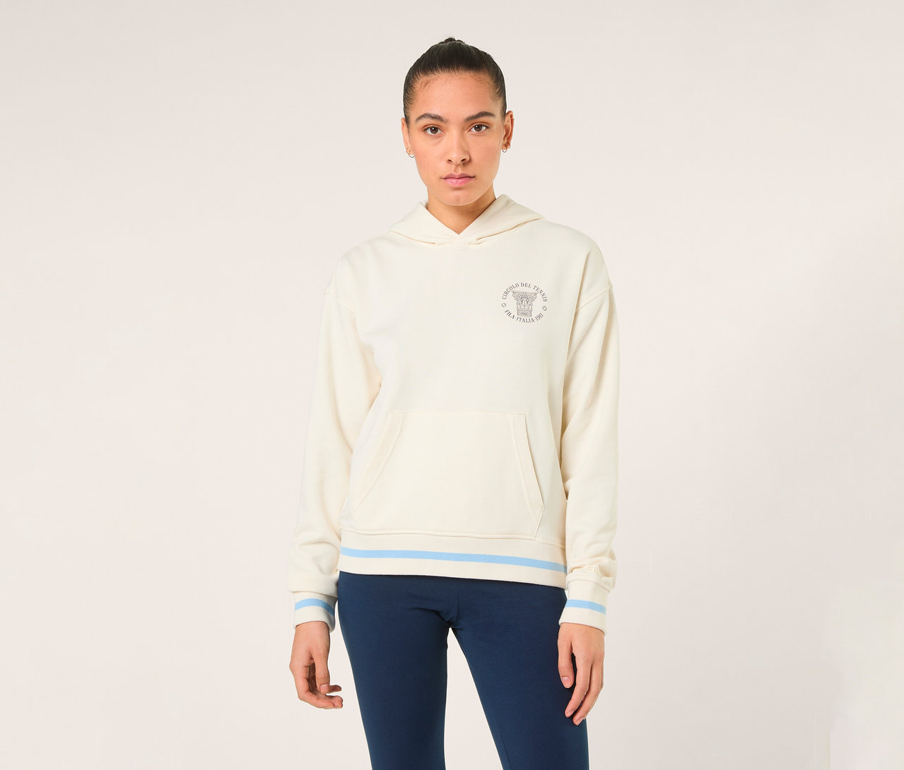 Eine Frau trägt einen off-white FILA Damen-Hoodie „MARLIA“ mit blauen Streifen an Saum und Ärmelbündchen sowie einem Logo auf der Brust.