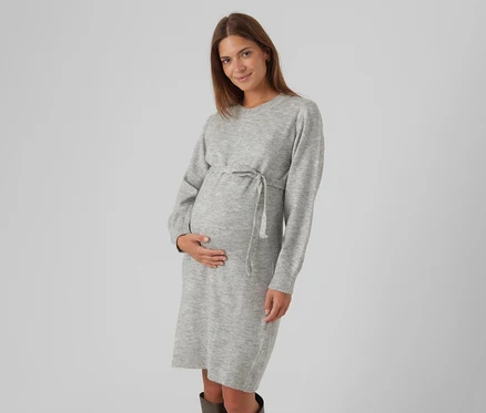Schwangere Frau mit langen braunen Haaren, die ein hellgraues Strickkleid mit Kordelzug an der Taille trägt, ihren Babybauch sanft hält und lächelt.
