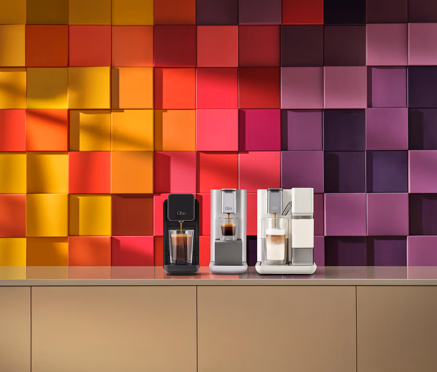 Drei Qbo Kaffeemaschinen auf einer beigen Arbeitsplatte. Eine schwarze Maschine gießt Espresso in ein Glas, eine silberne Maschine gießt Espresso in ein weiteres Glas und eine weiße Maschine mit Milchaufschäumer bereitet einen Latte zu. Dahinter befindet sich eine Wand mit 3D-Quadratfliesen in einem Farbverlauf von Gelb zu Violett.