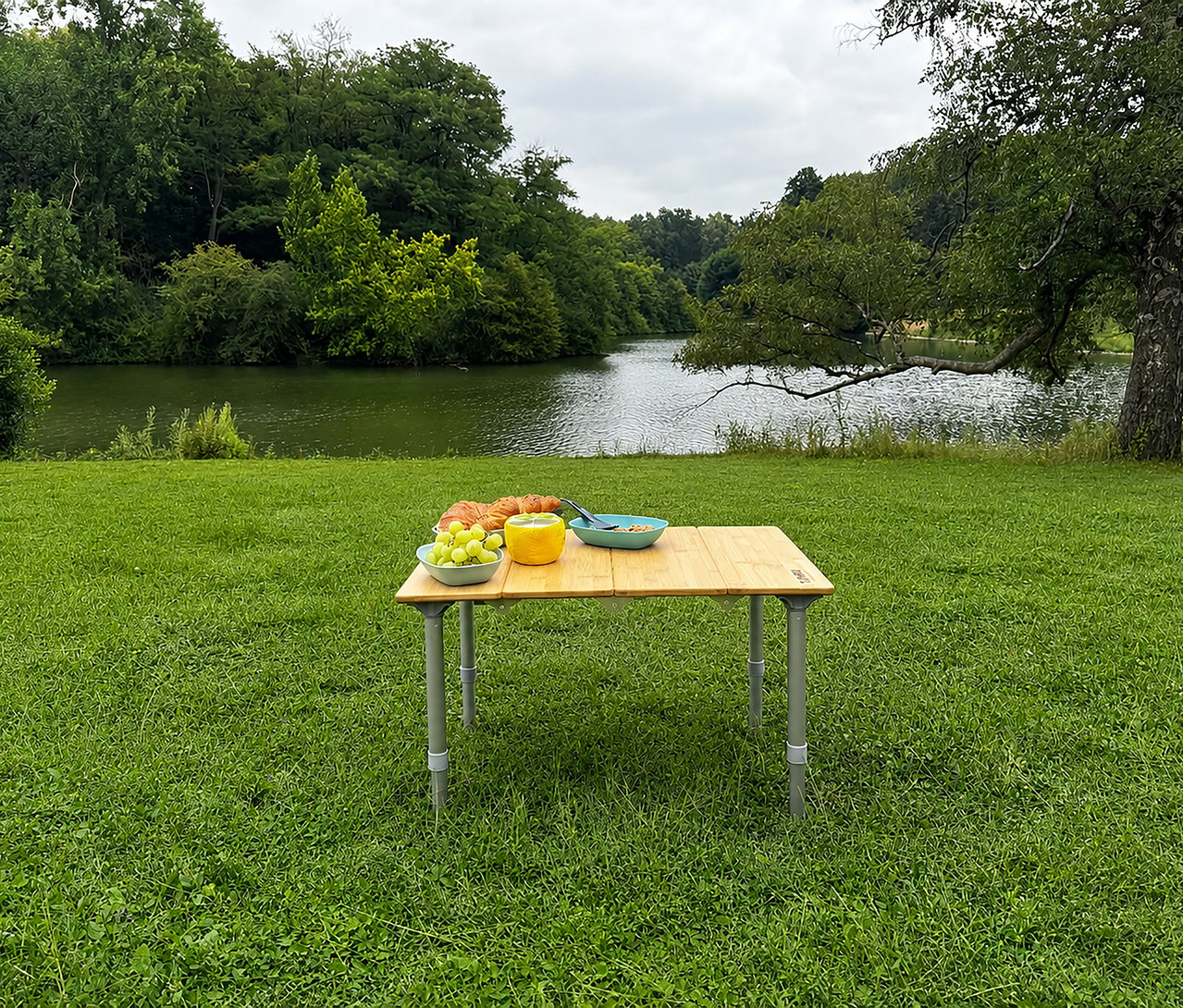 Ein Tambu Campingtisch »TEBO« steht auf grünem Gras am See, beladen mit Frühstück wie Trauben, Croissants, einem gelben Becher und einer blauen Schale.