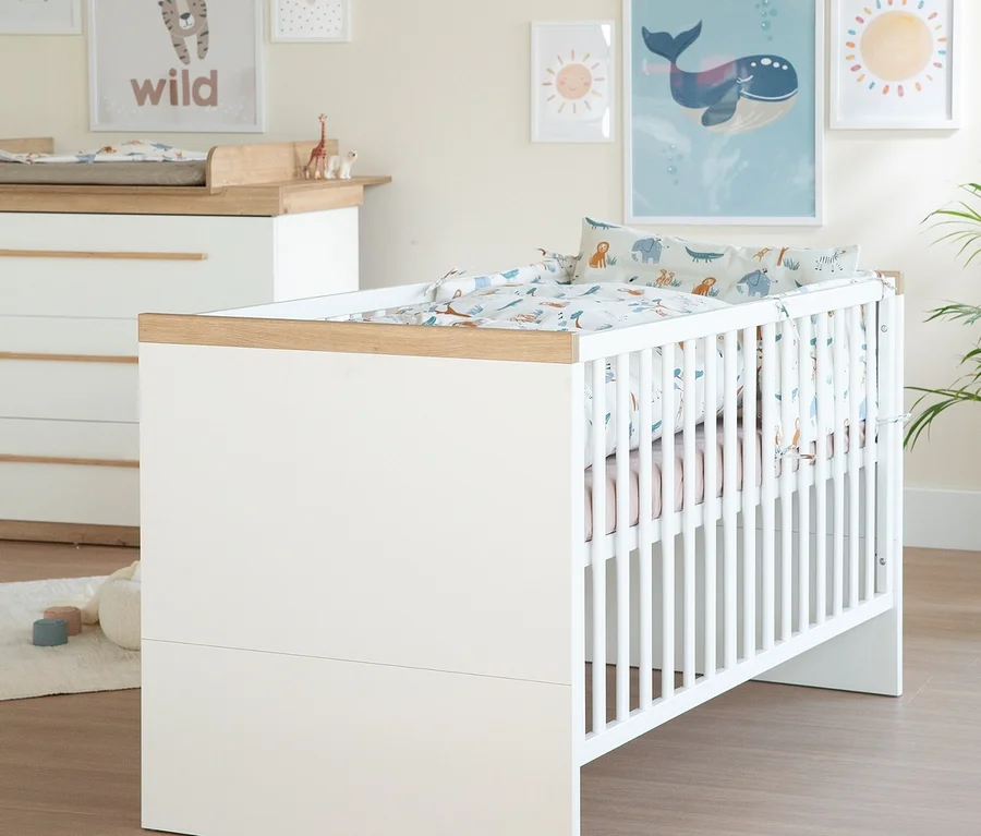 Ein Kinderzimmer mit einem weißen Gitterbett und einer passenden Wickelkommode aus dem Roba 3-teiligen Kinderzimmer-Set »Lumo«. Beide Möbelstücke haben Holzdetails. Das Gitterbett ist mit weißer Bettwäsche mit Tiermotiven bezogen, die Wickelkommode hat eine Wickelauflage.