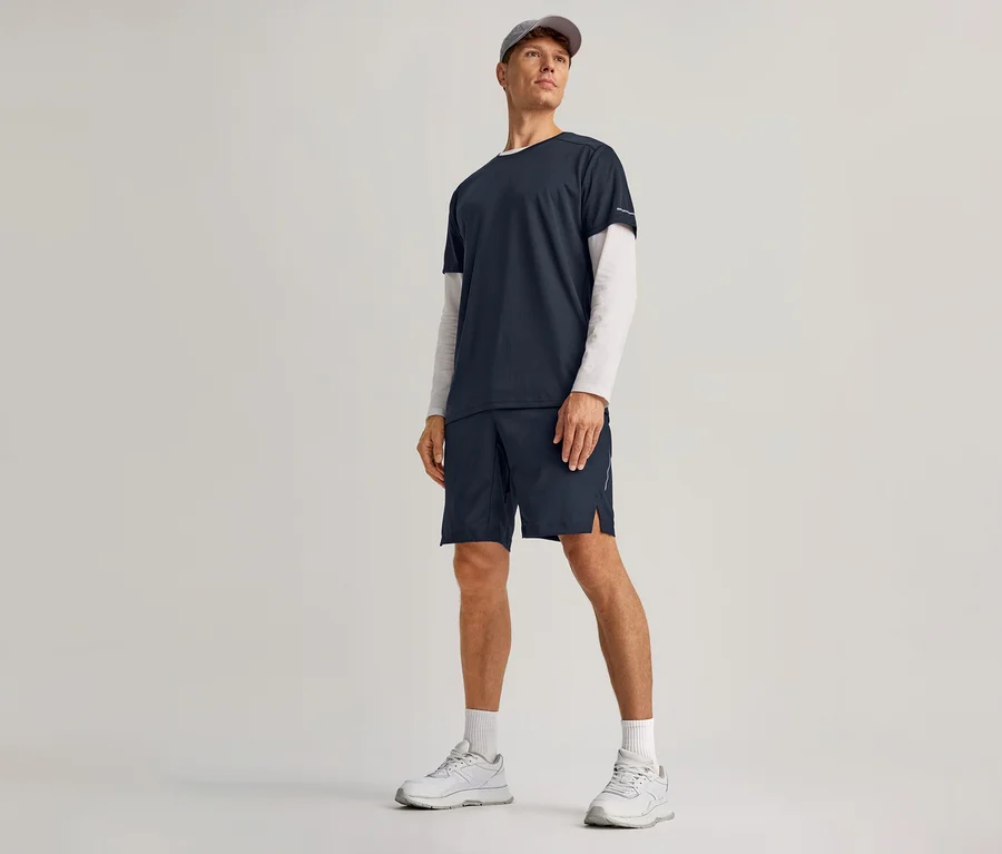 Mann trägt navy Sportshorts, ein navy T-Shirt über einem weißen Langarmshirt, weiße Socken, weiße Turnschuhe und eine graue Kappe.