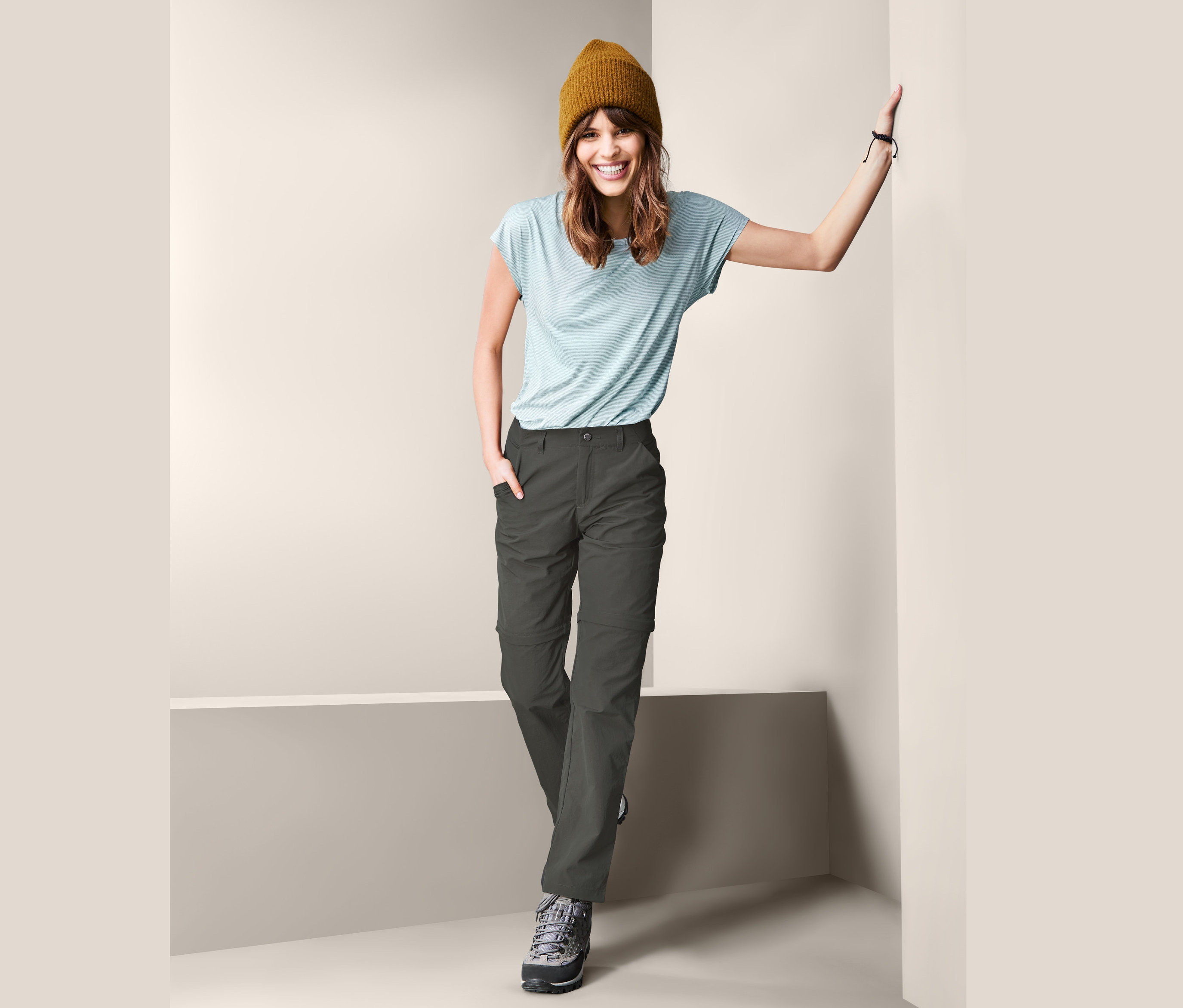 Lächelnde Frau posiert mit brauner Mütze, hellblauem T-Shirt und abzippbarer Funktionshose.