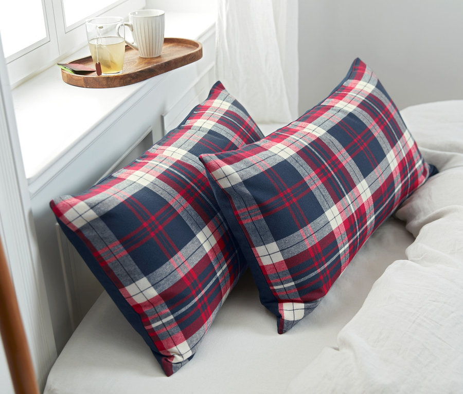 Zwei Flanell-Kopfkissenbezüge mit rot-weiß-blauem Karomuster liegen auf einem Bett. Daneben liegen eine graue Decke und ein Tablett mit Tassen.