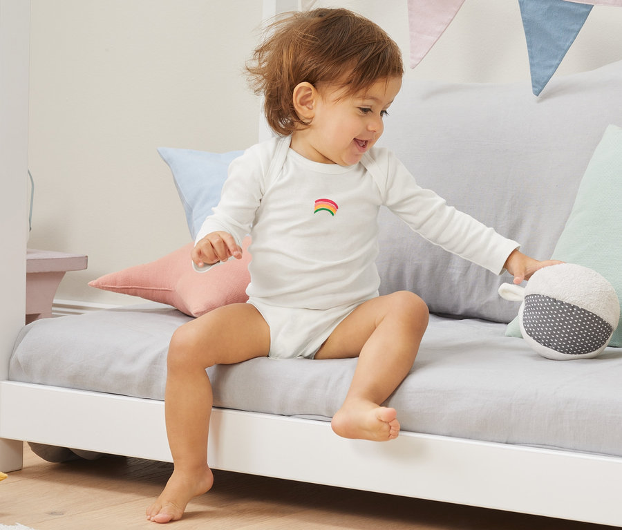 Ein Kind sitzt auf einem Bett und spielt mit einem Spielzeug, es trägt einen 5er Langarm-Body aus Bio-Baumwolle.