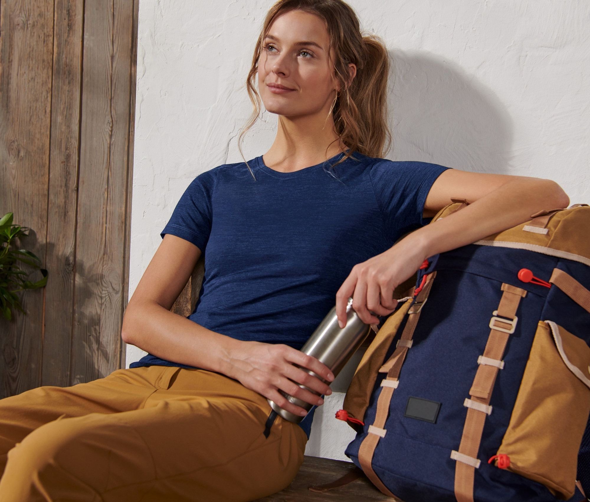 Frau sitzt mit Rucksack, hält eine Wasserflasche und trägt ein Funktionsshirt mit Wolle (Merinowolle).