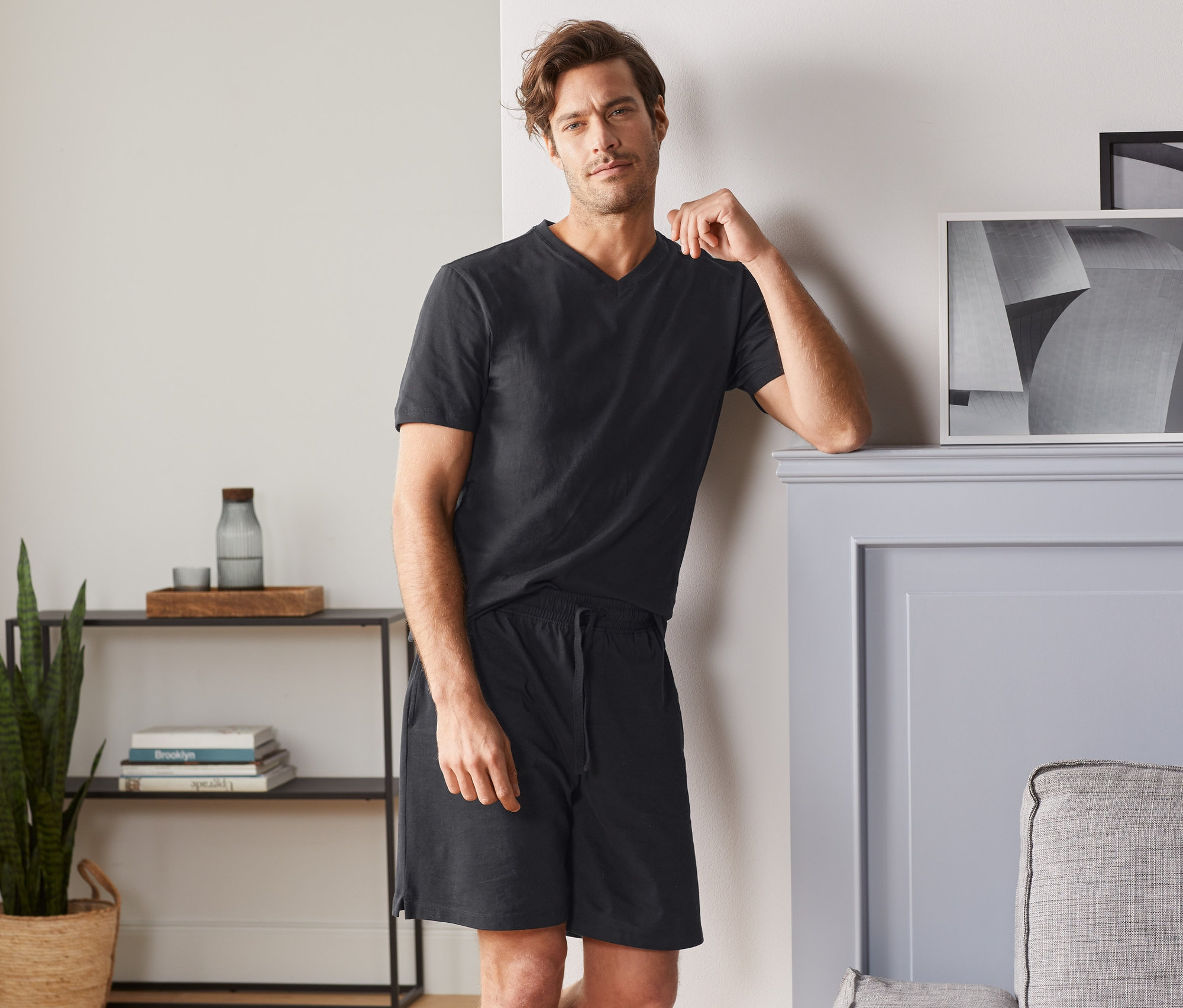 Mann an eine Wand gelehnt, trägt schwarzes T-Shirt mit V-Ausschnitt und schwarze Shorts.