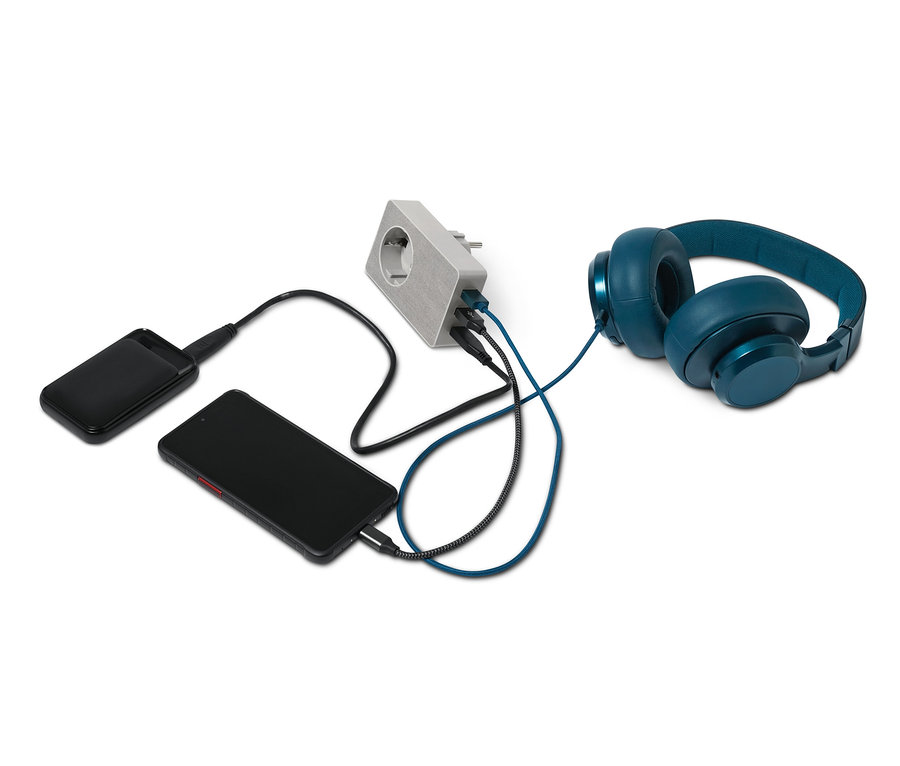 Ein schwarzes Smartphone und eine Powerbank werden über einen 4-in-1-USB-Steckdosen-Adapter aufgeladen. Blaue Kopfhörer liegen neben dem Adapter.