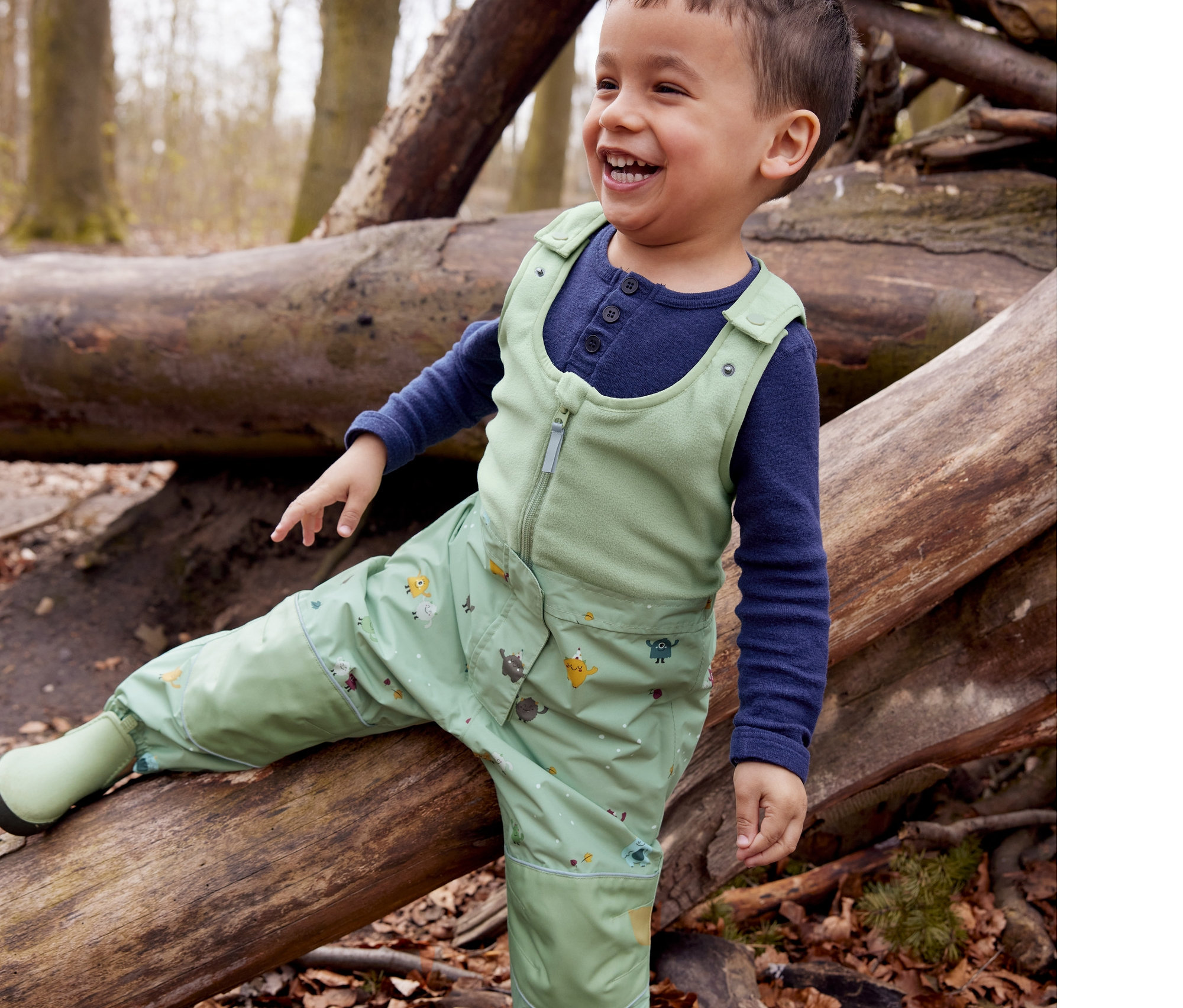 Ein lächelnder kleiner Junge sitzt auf einem Baumstamm in seinen Chelseaboot-Gummistiefeln und seiner Thermo-Regenhose.