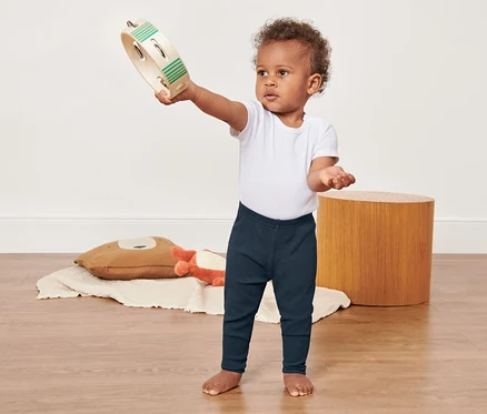 Ein lockiges Baby in einem weißen T-Shirt und 3 Baby-und-Kleinkinder-Leggings hält ein Tamburin hoch.