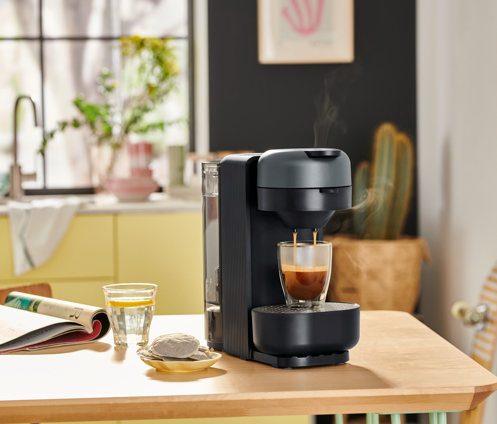 »Call me Pad« Kaffee Padmaschine Graphite befüllt eine Tasse mit Kaffee auf einem Tisch mit einer Zeitung und einem Glas Wasser.