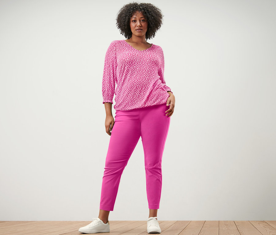 Frau posiert in pinkem, bedrucktem Blusenshirt und pinker Stretchhose.