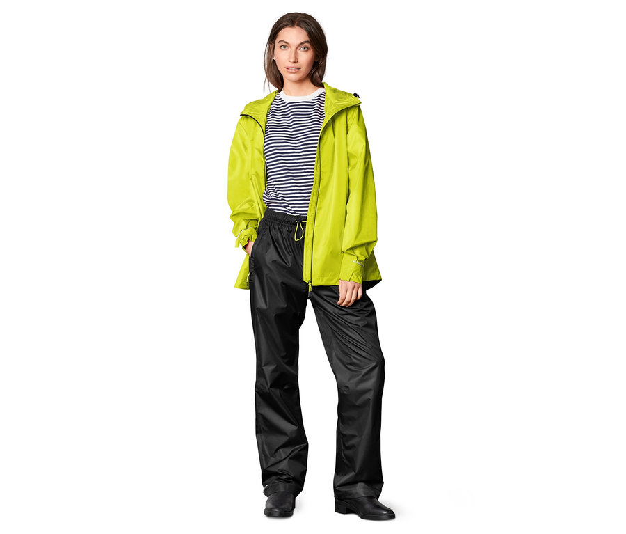 Eine Frau steht vor einem weißen Hintergrund und trägt eine schwarze Regenhose und eine gelbe Jacke.