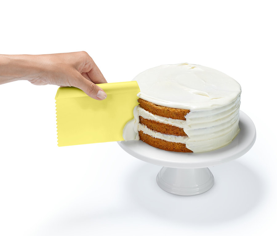 Eine Hand verwendet einen gelben Schaber aus einem Backhelfer-Set, um einem dreischichtigen Kuchen mit weißem Guss auf einem weißen Sockel zu versehen.