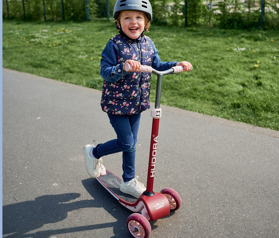 Ein Kind mit Helm fährt mit einem bordeauxroten HUDORA Tri-Scooter auf dem Gehweg.
