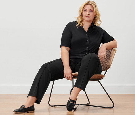 Frau sitzt auf einem Stuhl in schwarzem Blusenshirt mit Knopfleiste, schwarzer Sweathose und Leder-Loafern.