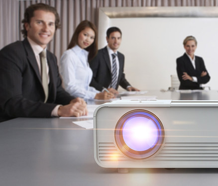 Weißer Mini-LED-HD-Beamer auf einem Tisch, im Hintergrund verschwommene Personen.