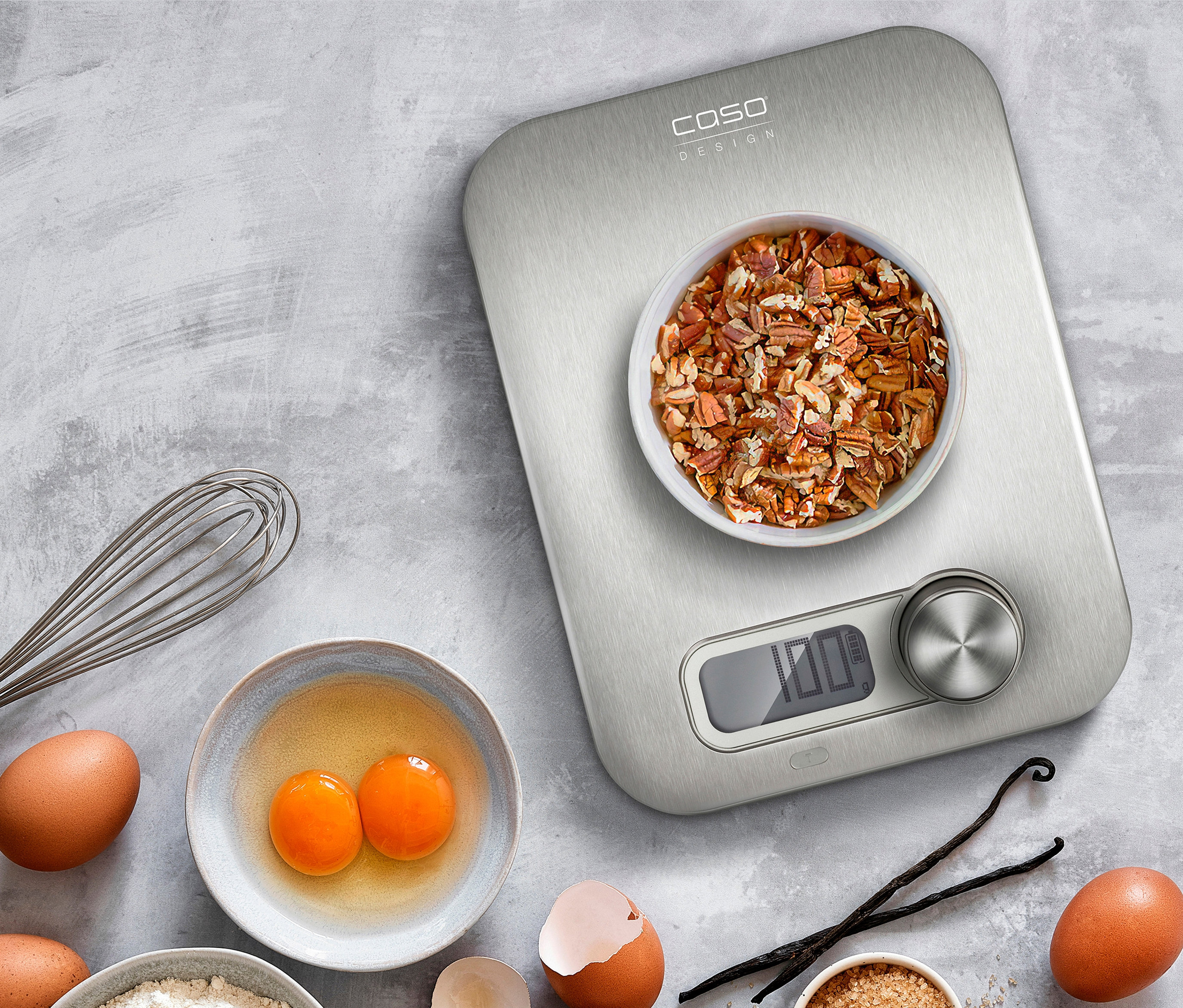 CASO Küchenwaage »Kitchen Energy« mit Nüssen in einer Schüssel, Eiern, Mehl, Zucker und Honig auf grauem Hintergrund.