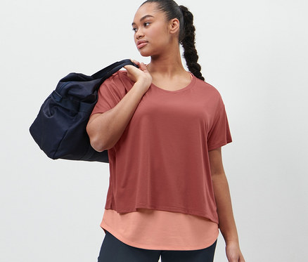Frau mit dunkelblauer Sporttasche über der Schulter trägt ein terrakottafarbenes 2-in-1-Kurzarm-Sportshirt und eine Bodyforming-Sporthose.